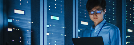 An IT technician in glasses works on a laptop in a blue-lit server room